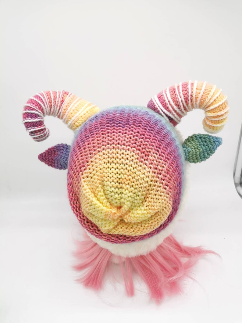 Puede incluir: Un gorro de punto arco&iacute;ris con rayas blancas y rosas en los cuernos. El gorro est&aacute; en una peluca rosa.
