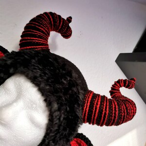 K&ouml;nnte beinhalten: Ein schwarzes Kunstfell-Stirnband mit zwei gebogenen H&ouml;rnern. Die H&ouml;rner sind schwarz mit roten Streifen. Rote geh&auml;kelte Ohren sind am Stirnband befestigt. Die H&ouml;rner sind ein einzigartiges und auff&auml;lliges Design.
