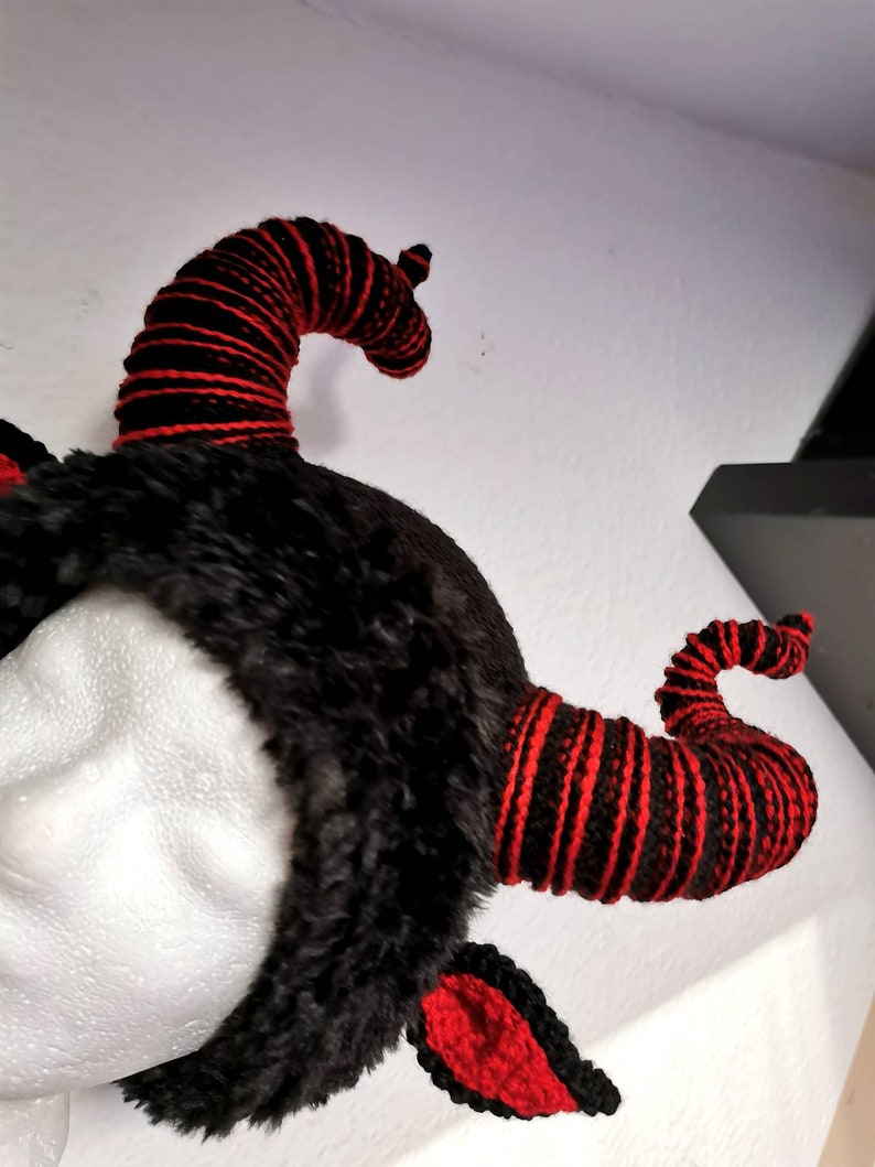 K&ouml;nnte beinhalten: Ein schwarzes Kunstfell-Stirnband mit zwei gebogenen H&ouml;rnern. Die H&ouml;rner sind schwarz mit roten Streifen. Rote geh&auml;kelte Ohren sind am Stirnband befestigt. Die H&ouml;rner sind ein einzigartiges und auff&auml;lliges Design.