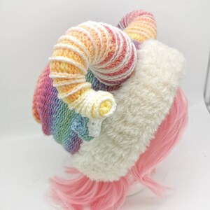 Puede incluir: Un gorro de crochet arco&iacute;ris con un borde de piel sint&eacute;tica blanca y dos grandes cuernos de crochet arco&iacute;ris rizados.