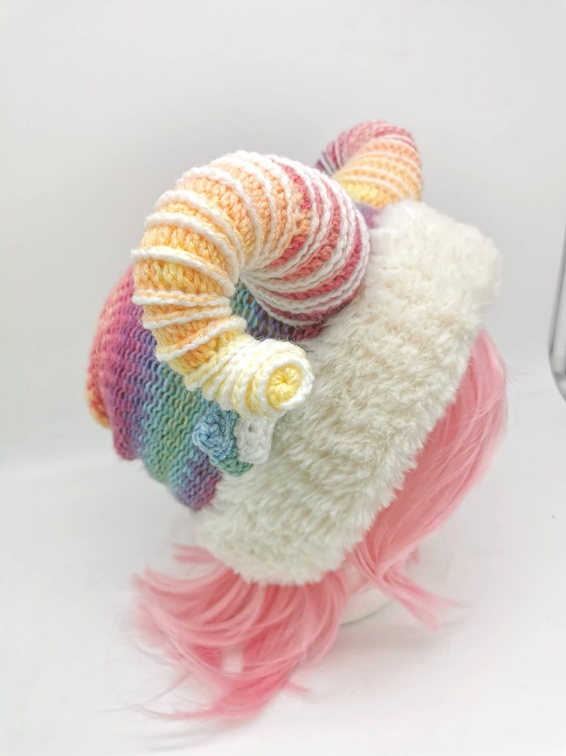 Puede incluir: Un gorro de crochet arco&iacute;ris con un borde de piel sint&eacute;tica blanca y dos grandes cuernos de crochet arco&iacute;ris rizados.