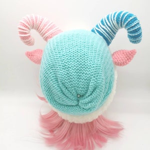 K&ouml;nnte beinhalten: Eine gestrickte M&uuml;tze mit rosa und blauen Zuckerstangenh&ouml;rnern, einem hellblauen K&ouml;rper und einem wei&szlig;en Pelzbesatz. Die M&uuml;tze sitzt auf einer rosafarbenen Per&uuml;cke.