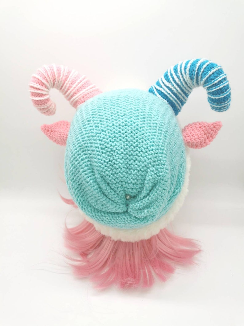 K&ouml;nnte beinhalten: Eine gestrickte M&uuml;tze mit rosa und blauen Zuckerstangenh&ouml;rnern, einem hellblauen K&ouml;rper und einem wei&szlig;en Pelzbesatz. Die M&uuml;tze sitzt auf einer rosafarbenen Per&uuml;cke.