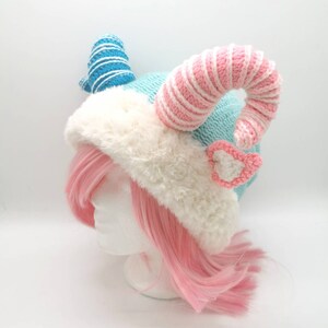 K&ouml;nnte beinhalten: Eine blaue und rosafarbene H&auml;kelm&uuml;tze mit wei&szlig;em Pelzbesatz und zwei gekr&auml;uselten H&ouml;rnern. Ein Horn ist blau-wei&szlig; gestreift, das andere rosa-wei&szlig; gestreift.