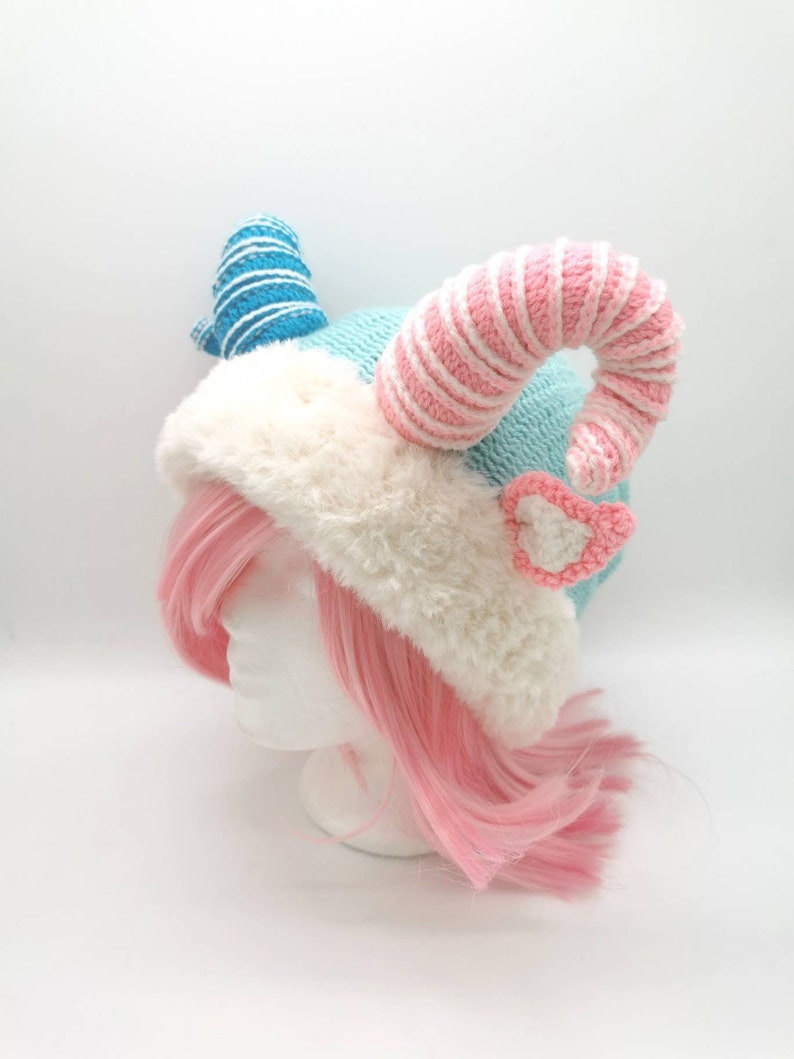 K&ouml;nnte beinhalten: Eine blaue und rosafarbene H&auml;kelm&uuml;tze mit wei&szlig;em Pelzbesatz und zwei gekr&auml;uselten H&ouml;rnern. Ein Horn ist blau-wei&szlig; gestreift, das andere rosa-wei&szlig; gestreift.