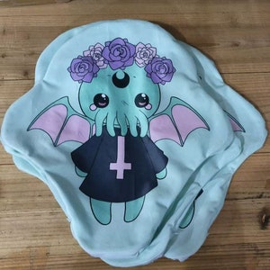 May include: A light blue pillow with a graphic print of a green, tentacled creature wearing a black robe with a cross. The creature has large eyes and is surrounded by pink flowers.