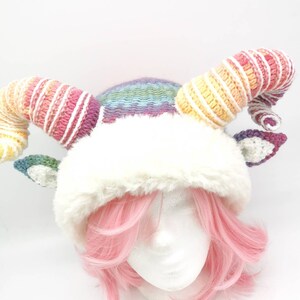 Puede incluir: Un gorro de crochet arco&iacute;ris con un borde de piel sint&eacute;tica blanca y dos cuernos largos de arco&iacute;ris enroscados. El gorro se lleva en una cabeza de maniqu&iacute; blanca con pelo rosa.