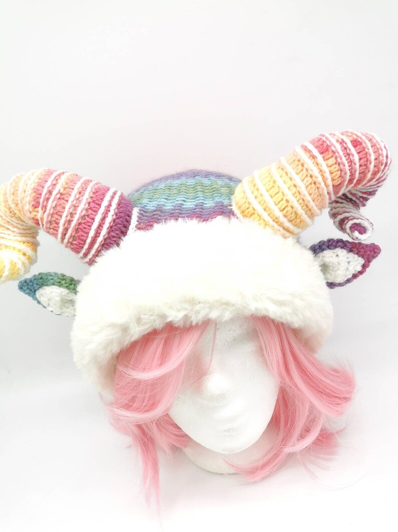 Puede incluir: Un gorro de crochet arco&iacute;ris con un borde de piel sint&eacute;tica blanca y dos cuernos largos de arco&iacute;ris enroscados. El gorro se lleva en una cabeza de maniqu&iacute; blanca con pelo rosa.