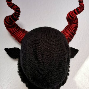 K&ouml;nnte beinhalten: Eine schwarze Strickm&uuml;tze mit zwei gro&szlig;en, gebogenen H&ouml;rnern. Die H&ouml;rner sind rot mit schwarzen Streifen. Die M&uuml;tze hat auch kleine, schwarze, ohr&auml;hnliche Klappen. Die M&uuml;tze wird vor einem wei&szlig;en Hintergrund pr&auml;sentiert.