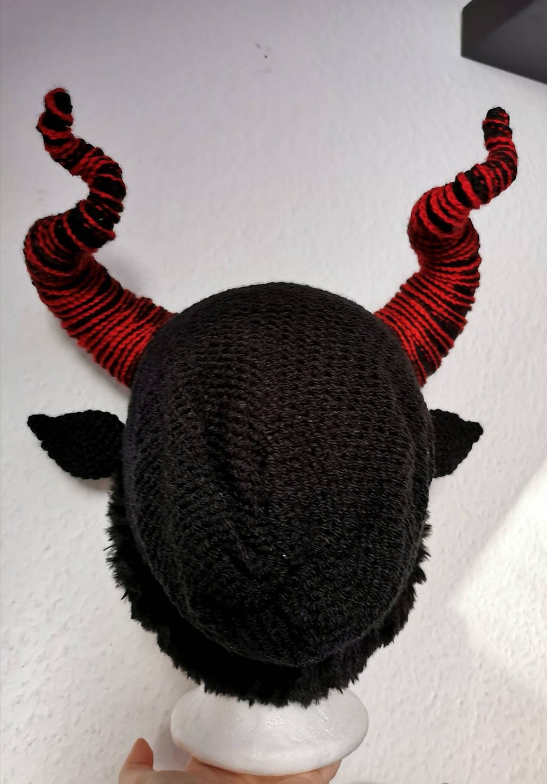 K&ouml;nnte beinhalten: Eine schwarze Strickm&uuml;tze mit zwei gro&szlig;en, gebogenen H&ouml;rnern. Die H&ouml;rner sind rot mit schwarzen Streifen. Die M&uuml;tze hat auch kleine, schwarze, ohr&auml;hnliche Klappen. Die M&uuml;tze wird vor einem wei&szlig;en Hintergrund pr&auml;sentiert.