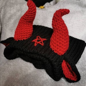 Puede incluir: Un gorro negro de punto con cuernos rojos de ganchillo y un diseño de pentagrama.