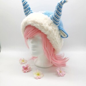 May include: A blue and white knitted hat with white fur trim and two long blue and white striped horns. The hat is on a white mannequin head with pink hair.