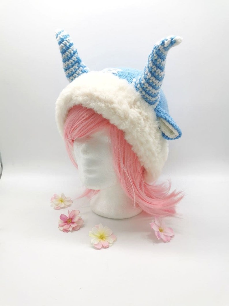 May include: A blue and white knitted hat with white fur trim and two long blue and white striped horns. The hat is on a white mannequin head with pink hair.