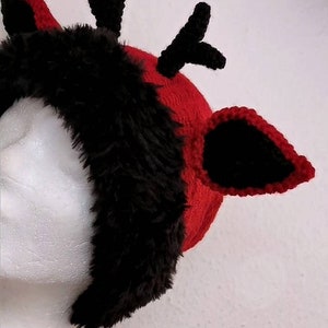 Puede incluir: Una diadema de cuernos de reno rojos y negros. La diadema presenta un gorro de punto rojo con ribete de piel sint&eacute;tica negra y cuernos y orejas de ganchillo negros. Las orejas est&aacute;n delineadas con ganchillo negro. La diadema est&aacute; sobre una cabeza de maniqu&iacute; blanca.