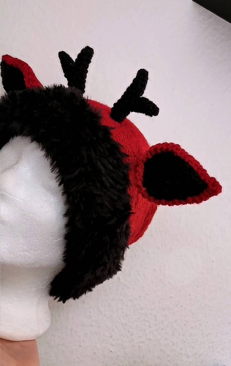 Puede incluir: Una diadema de cuernos de reno rojos y negros. La diadema presenta un gorro de punto rojo con ribete de piel sint&eacute;tica negra y cuernos y orejas de ganchillo negros. Las orejas est&aacute;n delineadas con ganchillo negro. La diadema est&aacute; sobre una cabeza de maniqu&iacute; blanca.