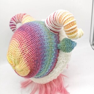 Puede incluir: Un animal de peluche de crochet arco&iacute;ris con pelaje blanco y pelo rosa. El animal tiene dos cuernos grandes y rizados.