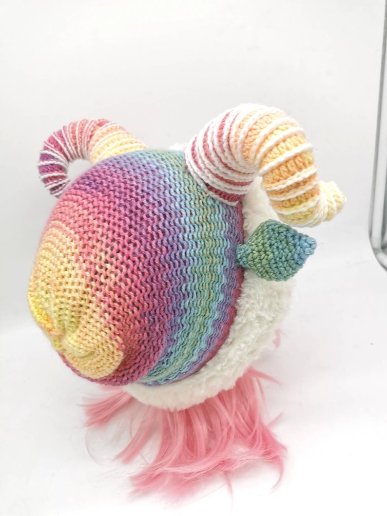 Puede incluir: Un animal de peluche de crochet arco&iacute;ris con pelaje blanco y pelo rosa. El animal tiene dos cuernos grandes y rizados.