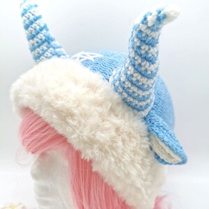 May include: A blue and white knitted hat with a fluffy white trim and two long, curled horns. The hat is worn on a mannequin with pink hair.