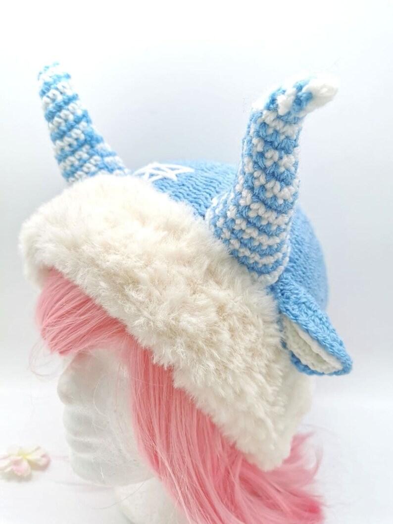 May include: A blue and white knitted hat with a fluffy white trim and two long, curled horns. The hat is worn on a mannequin with pink hair.
