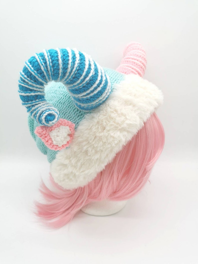 K&ouml;nnte beinhalten: Eine geh&auml;kelte M&uuml;tze mit einem blau-wei&szlig; gestreiften Horn, einer rosa-wei&szlig;en Blume und einem wei&szlig;en Pelzrand. Die M&uuml;tze sitzt auf einer rosafarbenen Per&uuml;cke.