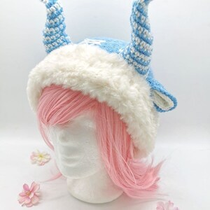 May include: A blue and white crocheted hat with white fur trim and two long, curled horns. The hat is on a white mannequin head with pink hair.