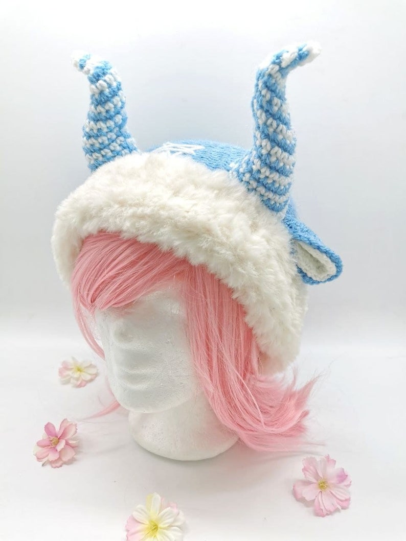 May include: A blue and white crocheted hat with white fur trim and two long, curled horns. The hat is on a white mannequin head with pink hair.