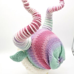 May include: A whimsical knit hat in a gradient of pink, purple, and green. The hat features two spiraling horns with alternating stripes of pink and white. The base of the hat has a soft, white trim and two green leaf-shaped accents.