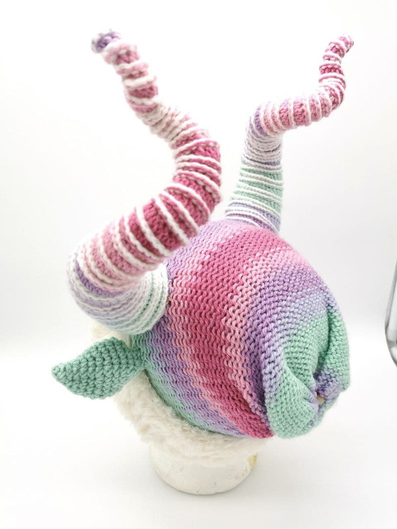 May include: A whimsical knit hat in a gradient of pink, purple, and green. The hat features two spiraling horns with alternating stripes of pink and white. The base of the hat has a soft, white trim and two green leaf-shaped accents.
