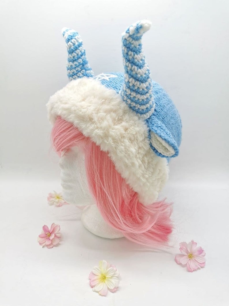May include: A blue and white crocheted hat with horns, a white faux fur brim, and a pink wig on a white mannequin head.