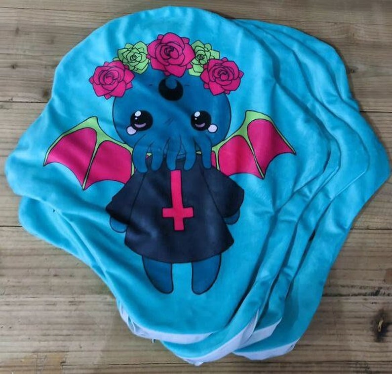 May include: A stack of blue fabric squares with a cartoon image of a Cthulhu-like creature wearing a black robe with a pentagram. The creature has a pink and green flower crown and is surrounded by pink and green wings.