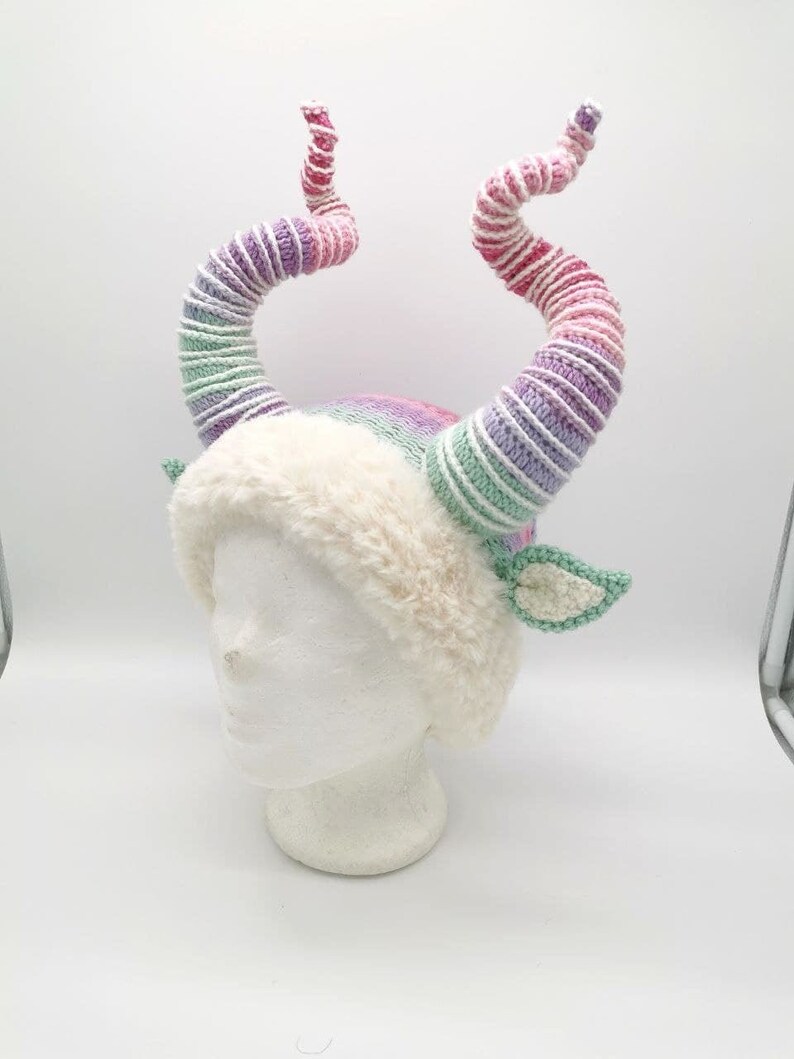 May include: A whimsical, handmade hat with large, spiraling horns in shades of pink, purple, and green. The hat features a fluffy, cream-colored brim and small, green crocheted leaf accents, creating a fantasy-themed accessory.