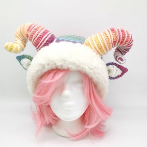 Puede incluir: Un gorro blanco de ganchillo con cuernos de colores arco&iacute;ris. El gorro tiene un borde de piel sint&eacute;tica blanca y se lleva sobre una peluca rosa.