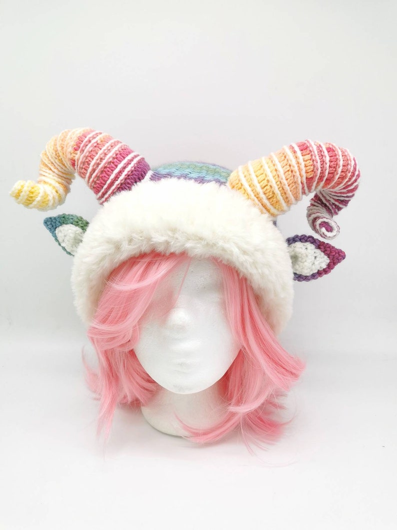 Puede incluir: Un gorro blanco de ganchillo con cuernos de colores arco&iacute;ris. El gorro tiene un borde de piel sint&eacute;tica blanca y se lleva sobre una peluca rosa.