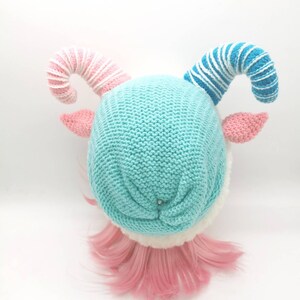 K&ouml;nnte beinhalten: Eine blaue Strickm&uuml;tze mit wei&szlig;em Pelzbesatz und zwei gekr&auml;uselten H&ouml;rnern, eines rosa-wei&szlig; gestreift und das andere blau-wei&szlig; gestreift. Die M&uuml;tze sitzt auf einem Puppenkopf mit rosa Haaren.