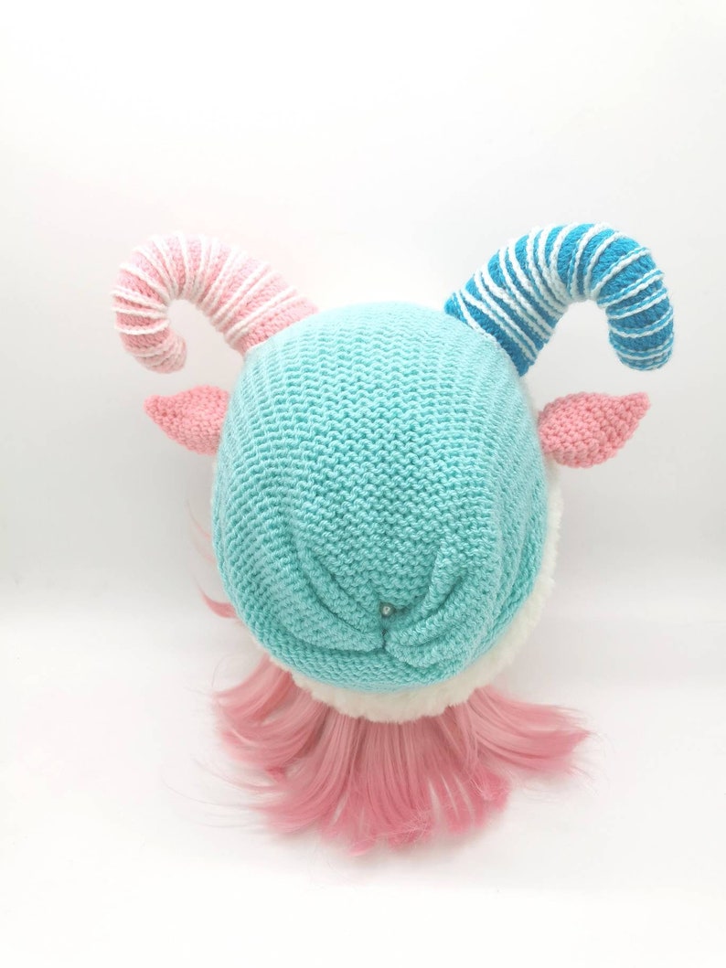 K&ouml;nnte beinhalten: Eine blaue Strickm&uuml;tze mit wei&szlig;em Pelzbesatz und zwei gekr&auml;uselten H&ouml;rnern, eines rosa-wei&szlig; gestreift und das andere blau-wei&szlig; gestreift. Die M&uuml;tze sitzt auf einem Puppenkopf mit rosa Haaren.