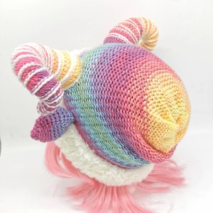 Puede incluir: Un gorro de punto de arco&iacute;ris con un borde blanco esponjoso y dos cuernos de hilo de arco&iacute;ris enroscados. El gorro est&aacute; en una peluca rosa.