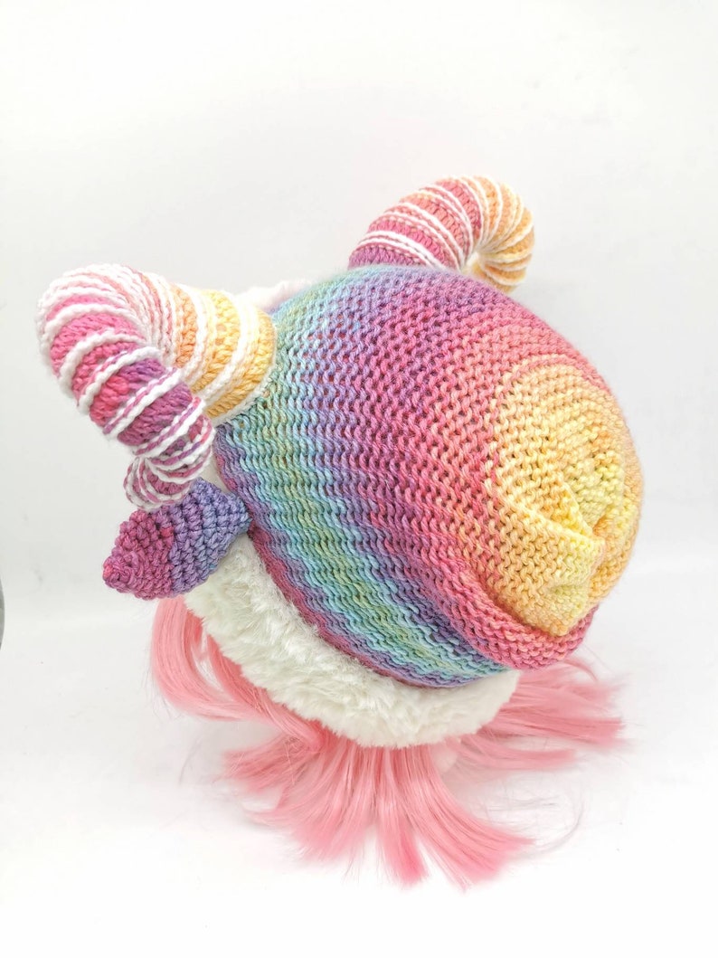 Puede incluir: Un gorro de punto de arco&iacute;ris con un borde blanco esponjoso y dos cuernos de hilo de arco&iacute;ris enroscados. El gorro est&aacute; en una peluca rosa.
