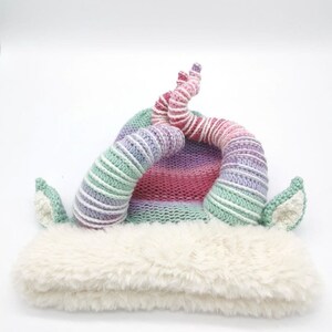 May include: A whimsical knit hat with a fluffy white brim and colorful horns. The hat features a striped pattern in shades of green, purple, pink, and white. The horns are curved and add a playful touch to the design.