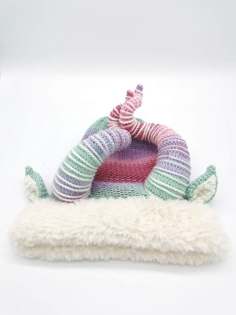 May include: A whimsical knit hat with a fluffy white brim and colorful horns. The hat features a striped pattern in shades of green, purple, pink, and white. The horns are curved and add a playful touch to the design.