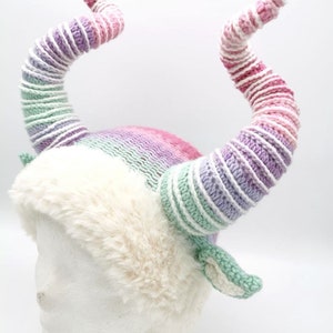 May include: A whimsical, handmade hat featuring large, curved horns in shades of pink, purple, and white. The hat has a fluffy, cream-colored faux fur trim and a knitted crown. The horns are approximately 12 inches long.