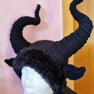 K&ouml;nnte beinhalten: Ein schwarzer Strickhut mit gro&szlig;en, gebogenen H&ouml;rnern und Kunstfellbesatz. Die H&ouml;rner sind ein markantes Merkmal, das sich von der Krone des Hutes nach oben windet. Der Hut ist so gestaltet, dass er einem geh&ouml;rnten Wesen &auml;hnelt.