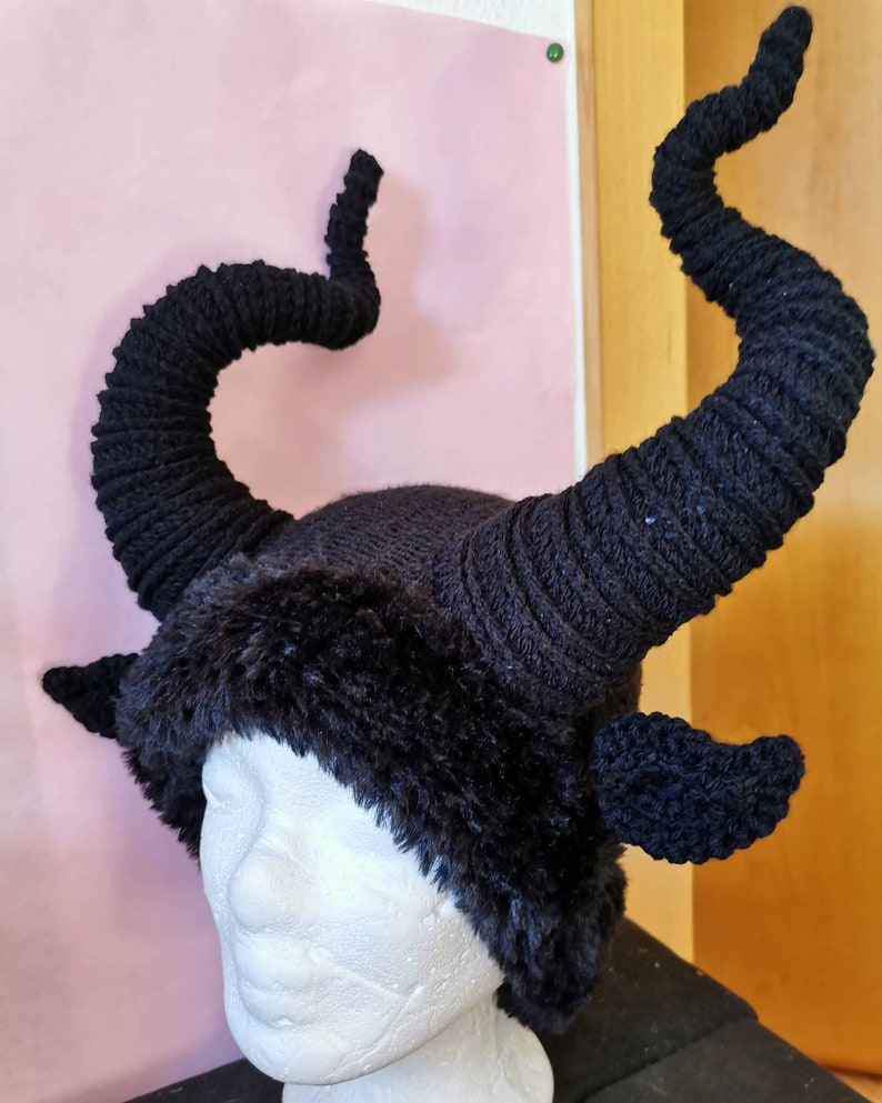 K&ouml;nnte beinhalten: Ein schwarzer Strickhut mit gro&szlig;en, gebogenen H&ouml;rnern und Kunstfellbesatz. Die H&ouml;rner sind ein markantes Merkmal, das sich von der Krone des Hutes nach oben windet. Der Hut ist so gestaltet, dass er einem geh&ouml;rnten Wesen &auml;hnelt.