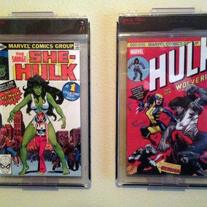 May include: Two comic books in protective plastic cases. The left comic book is green and features the She-Hulk character. The right comic book is red and features the Hulk and Wolverine characters. The text on the left comic book reads "The Savage She-Hulk" and "Stan Lee Presents The She-Hulk Lives!" The text on the right comic book reads "Marvel Comics Group Up" and "Hulk and Now Wolverine!"