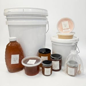 May include: A selection of honey products, including a large jug, jars, and tubs, with white labels featuring a bee graphic. The honey varies in colour from light amber to dark brown. White buckets are also present.