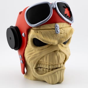 May include: A beige skull-shaped figurine wearing a red aviator helmet with black goggles. The helmet has black ear covers and silver accents. The skull has black eyes and a menacing expression. The figurine is on a white background.