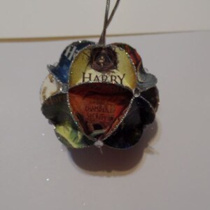 Puede incluir: Un adorno navideño plateado y dorado con un diseño de portada de libro de Harry Potter. El adorno presenta el texto "Harry Potter y la Cámara Secreta".