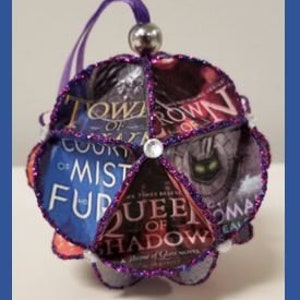 Könnte beinhalten: Ein lila und silberner Anhänger aus Buchseiten mit den Titeln "A Court of Mist and Fury", "Queen of Shadows", "A Court of Thorns and Roses" und "A Court of Wings and Ruin".