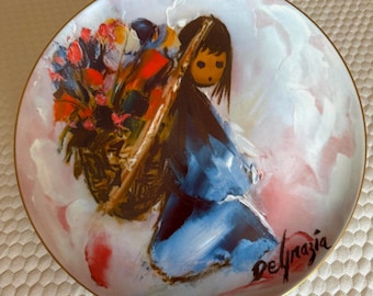 May include: A round decorative ceramic plate with a gold-tone rim featuring a painted illustration of a person with dark hair wearing a blue garment, carrying a large basket filled with colorful flowers, signed Delgrazia in the corner.