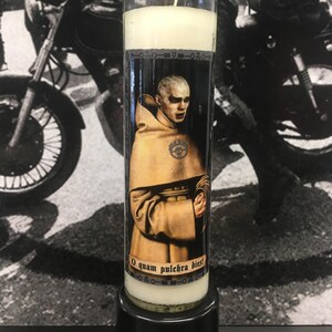 May include: A white candle in a glass jar with a black and white image of a man in a brown robe. The image has the text "O quam pulchra dies!" printed below it.