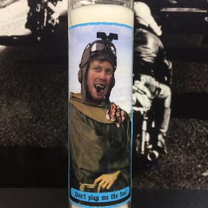 May include: A clear glass candle with a white wax interior. The candle features a label with a vintage image of a man in a pilot's helmet and the text "Don't play me the fool". The background is a black and white photo.